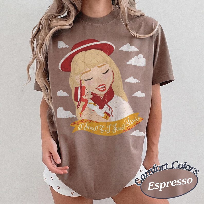 Pode incluir: Uma t-shirt castanha de manga curta com uma ilustra&ccedil;&atilde;o gr&aacute;fica de uma pessoa loira com um chap&eacute;u e um len&ccedil;o vermelhos, segurando um auscultador de telefone vermelho. Abaixo da figura, uma faixa amarela cont&eacute;m o texto I Knew It, I Knew You.