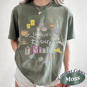 Op de afbeelding: Olijfgroen T-shirt met een kleurrijke Disney-thema afbeelding. Het ontwerp bevat een kasteel, lamp, pompoen en de tekst "Literally just a Disney Girl". Het shirt heeft een relaxte pasvorm en de woorden "Comfort Colors Moss" staan rechtsonder.