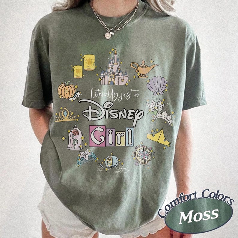 Op de afbeelding: Olijfgroen T-shirt met een kleurrijke Disney-thema afbeelding. Het ontwerp bevat een kasteel, lamp, pompoen en de tekst "Literally just a Disney Girl". Het shirt heeft een relaxte pasvorm en de woorden "Comfort Colors Moss" staan rechtsonder.