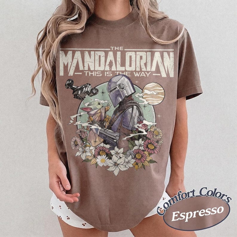 May include: A brown t-shirt featuring a graphic print of The Mandalorian and Grogu. The text reads THE MANDALORIAN THIS IS THE WAY. The design includes a spaceship, a planet, and a floral arrangement at the base. The shirt has a crew neckline.