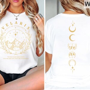 May include: White t-shirt with gold text and graphics. The front features "VELARIS CITY OF STARLIGHT" with a mountain and star design. The back displays a vertical design of moons, stars, and a globe. The shirt includes the text "TO THE STARS WHO LISTEN AND THE DREAMS THAT ARE ANSWERED."
