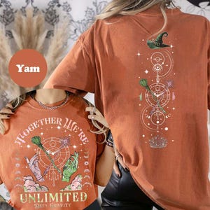 May include: Orange t-shirt with a graphic of a witch's hat, broomsticks, and stars. The text on the shirt says "Together We're Unlimited - Defy Gravity".