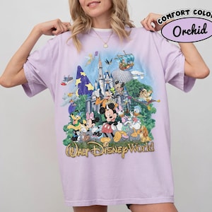 May include: A light purple short-sleeve t-shirt featuring a graphic print of Walt Disney World with various characters including Mickey Mouse, Minnie Mouse, Donald Duck, and Goofy in front of a castle. The text Walt Disney World is printed below.