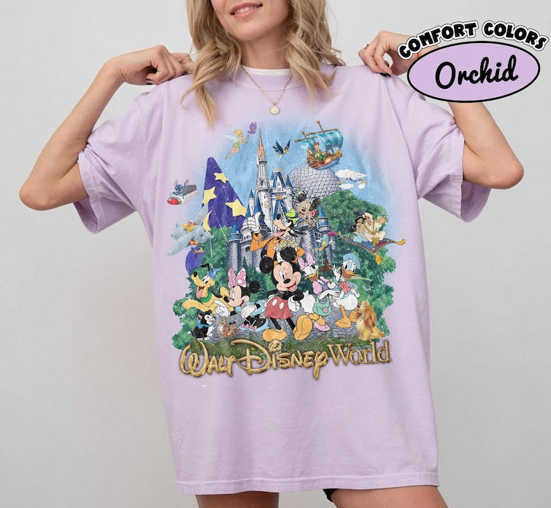 May include: A light purple short-sleeve t-shirt featuring a graphic print of Walt Disney World with various characters including Mickey Mouse, Minnie Mouse, Donald Duck, and Goofy in front of a castle. The text Walt Disney World is printed below.