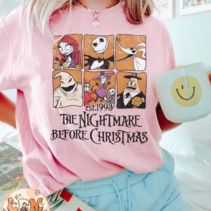 May include: A pink t-shirt with a vintage-style graphic of characters from the movie "The Nightmare Before Christmas". The graphic features Jack Skellington, Sally, Zero, Oogie Boogie, and other characters in a grid pattern. The text "est. 1993" and "The Nightmare Before Christmas" is printed below the graphic.