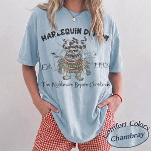 May include: A light blue short-sleeve t-shirt featuring a graphic of a mummy character from The Nightmare Before Christmas. The text reads HARLEQUIN DEMON, Est. 1993, and The Nightmare Before Christmas. The design includes spiderwebs and spiders.