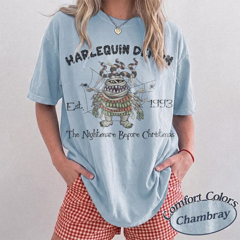 May include: A light blue short-sleeve t-shirt featuring a graphic of a mummy character from The Nightmare Before Christmas. The text reads HARLEQUIN DEMON, Est. 1993, and The Nightmare Before Christmas. The design includes spiderwebs and spiders.