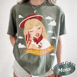 Pode incluir: Uma t-shirt de manga curta verde musgo com uma ilustra&ccedil;&atilde;o gr&aacute;fica de uma personagem loira a usar um chap&eacute;u e um cachecol vermelhos. A personagem segura uma pequena boneca. Abaixo da ilustra&ccedil;&atilde;o est&aacute; uma faixa com o texto I knew it, I knew you.