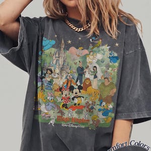 May include: A charcoal gray oversized t-shirt featuring a large, colorful graphic print of various animated characters, a castle, and the text Magic Kingdom Walt Disney World. The shirt includes a Comfort Colors Pepper label in the bottom corner.