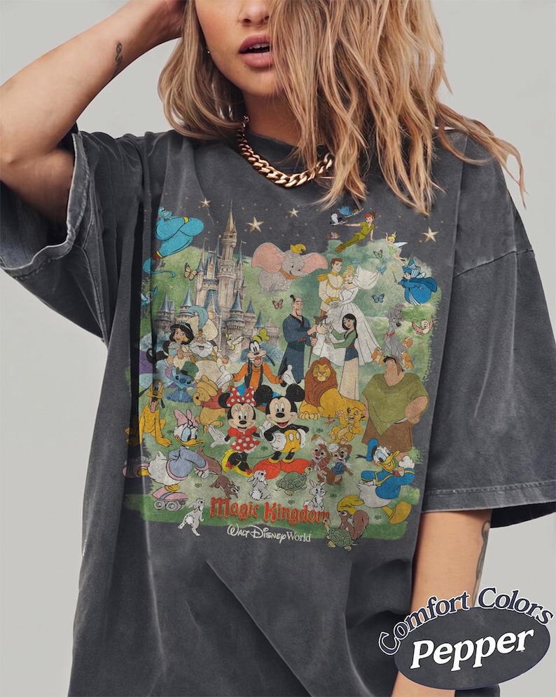 May include: A charcoal gray oversized t-shirt featuring a large, colorful graphic print of various animated characters, a castle, and the text Magic Kingdom Walt Disney World. The shirt includes a Comfort Colors Pepper label in the bottom corner.
