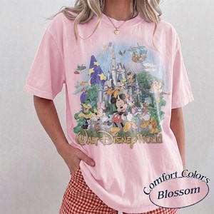 May include: A light pink short-sleeve t-shirt featuring a large, colorful graphic print of Walt Disney World with various cartoon characters, including Mickey Mouse, Minnie Mouse, Donald Duck, and Goofy, set against a castle background.