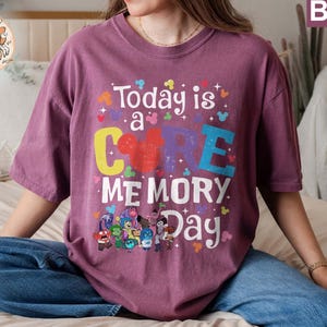 以下が含まれることがあります： 「Today is a CORE MEMORY Day」という文字と、漫画のミッキーマウスの頭が描かれた紫色のTシャツ。Tシャツには、映画「インサイド・ヘッド」の漫画のキャラクターのグループも描かれている。