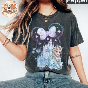 Puede incluir: Una camiseta gris oscuro con un dise&ntilde;o de acuarela que presenta un castillo, una princesa con cabello rubio y un lazo morado. El dise&ntilde;o est&aacute; rodeado por un halo azul claro y morado.