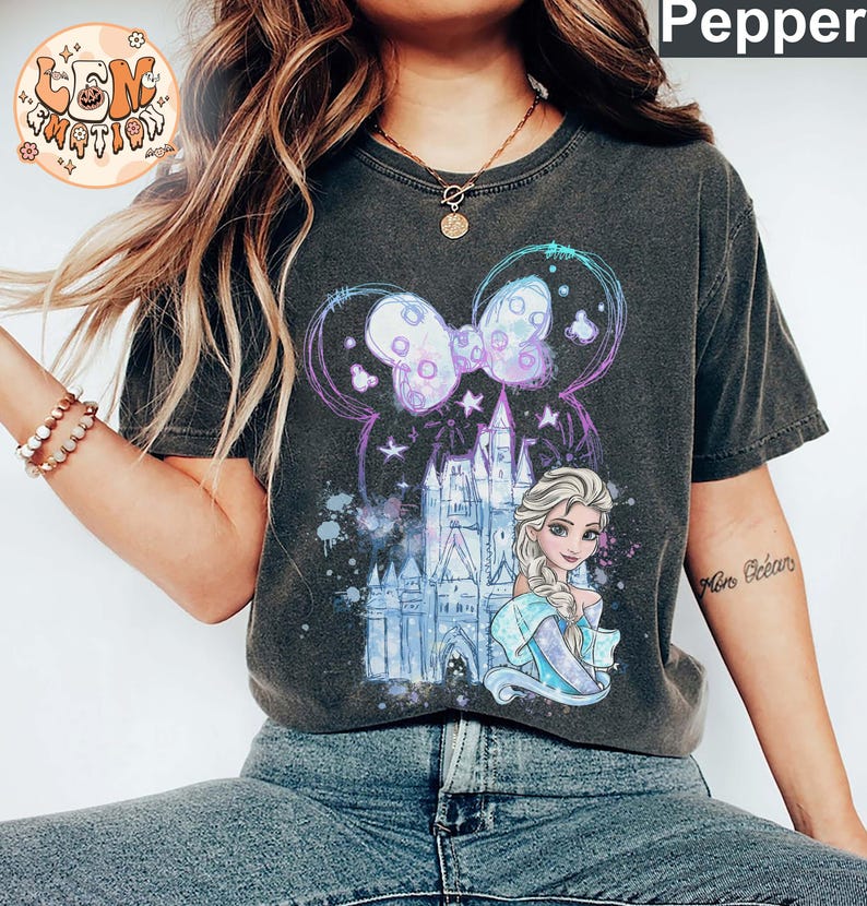 Puede incluir: Una camiseta gris oscuro con un dise&ntilde;o de acuarela que presenta un castillo, una princesa con cabello rubio y un lazo morado. El dise&ntilde;o est&aacute; rodeado por un halo azul claro y morado.