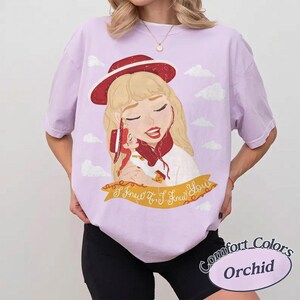 Pode incluir: Uma t-shirt roxo-claro com uma ilustra&ccedil;&atilde;o gr&aacute;fica de uma pessoa loira usando um chap&eacute;u e cachecol vermelhos, segurando uma boneca pequena. Abaixo da ilustra&ccedil;&atilde;o, h&aacute; uma faixa amarela com o texto I Knew It, I Knew You em letra cursiva.