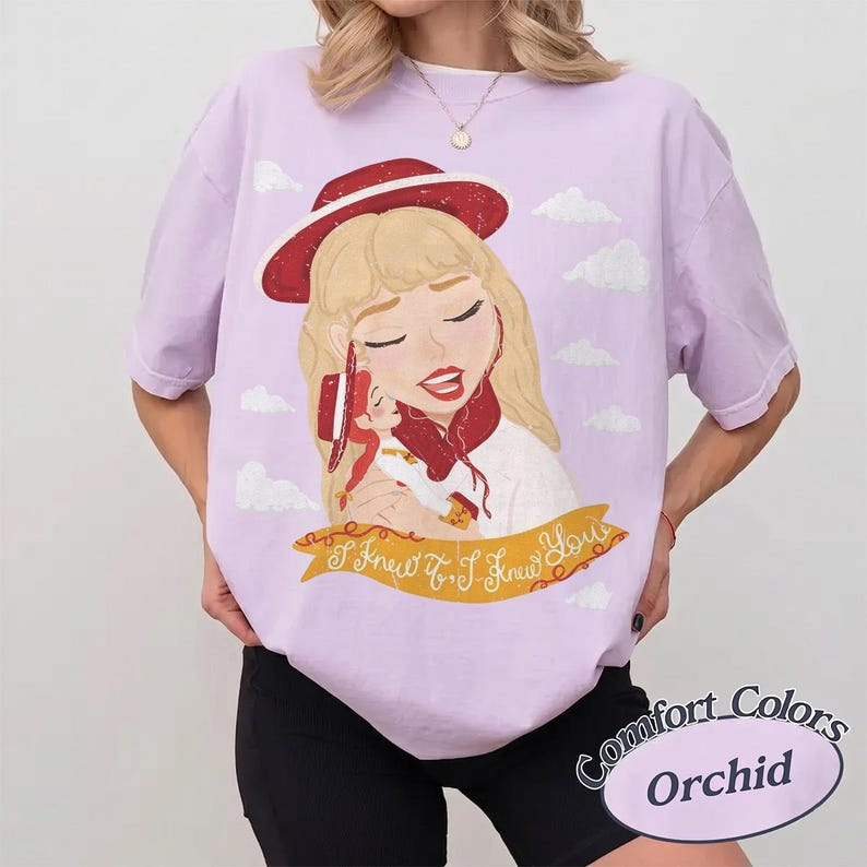 Pode incluir: Uma t-shirt roxo-claro com uma ilustra&ccedil;&atilde;o gr&aacute;fica de uma pessoa loira usando um chap&eacute;u e cachecol vermelhos, segurando uma boneca pequena. Abaixo da ilustra&ccedil;&atilde;o, h&aacute; uma faixa amarela com o texto I Knew It, I Knew You em letra cursiva.