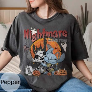 Puede incluir: Camiseta gris oscuro con un dise&ntilde;o tem&aacute;tico de Halloween. El dise&ntilde;o incluye las palabras "Nightmare on Main Street", personajes de dibujos animados, calabazas, una l&aacute;pida y una luna llena. La camiseta es de corte holgado.