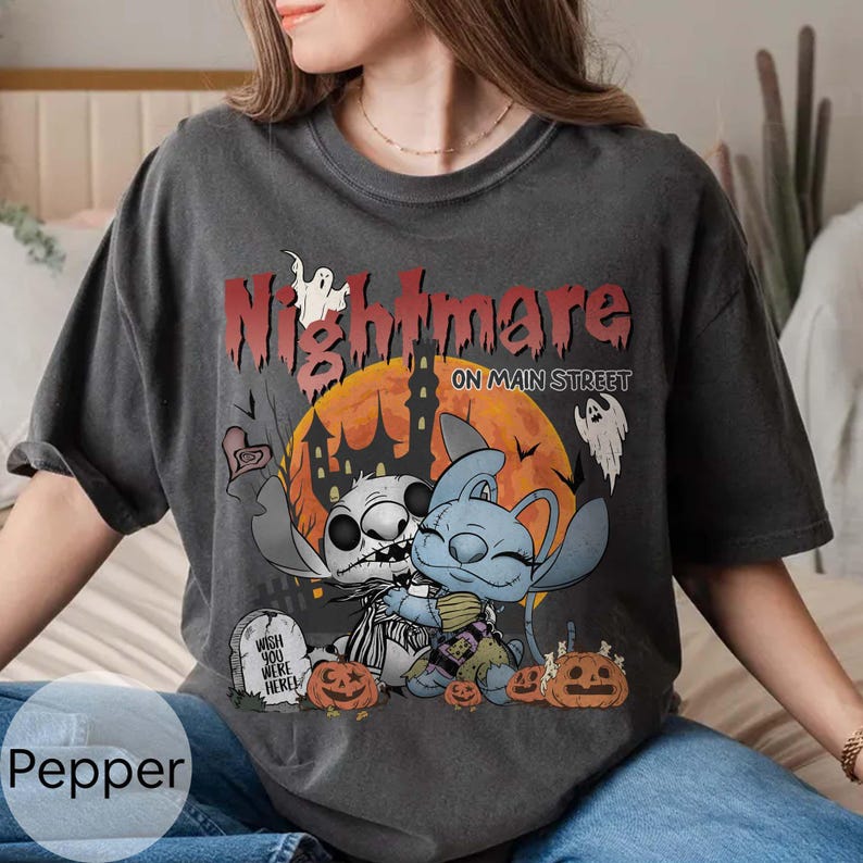 Puede incluir: Camiseta gris oscuro con un dise&ntilde;o tem&aacute;tico de Halloween. El dise&ntilde;o incluye las palabras "Nightmare on Main Street", personajes de dibujos animados, calabazas, una l&aacute;pida y una luna llena. La camiseta es de corte holgado.