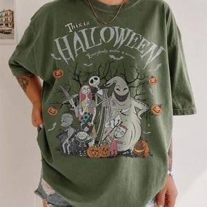 Pu&ograve; includere: T-shirt verde oliva con grafica di The Nightmare Before Christmas. Il design include Jack Skellington, Sally, Oogie Boogie e altri personaggi, con il testo "This is Halloween" e "Everybody make a scene". Sono presenti zucche arancioni.