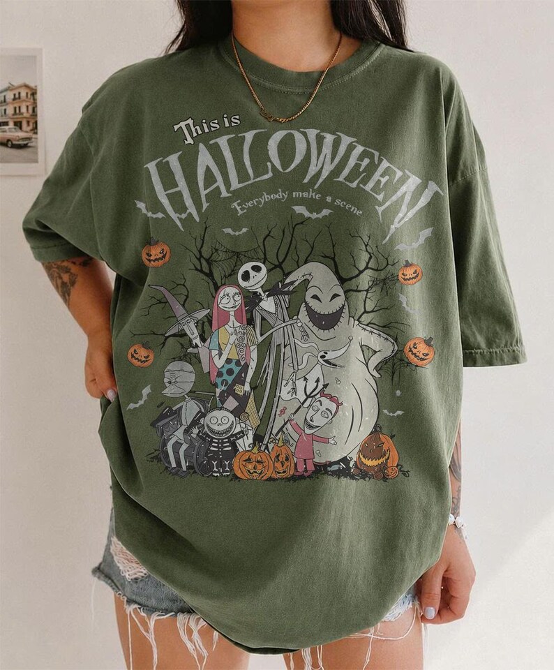 Pu&ograve; includere: T-shirt verde oliva con grafica di The Nightmare Before Christmas. Il design include Jack Skellington, Sally, Oogie Boogie e altri personaggi, con il testo "This is Halloween" e "Everybody make a scene". Sono presenti zucche arancioni.