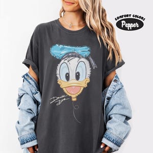 Pode incluir: Uma t-shirt de manga curta cinzento-carv&atilde;o com um gr&aacute;fico do rosto do Pato Donald com um la&ccedil;o azul, um bico amarelo e a assinatura Donald Duck escrita em branco. A t-shirt inclui uma etiqueta Comfort Colors Pepper no canto.