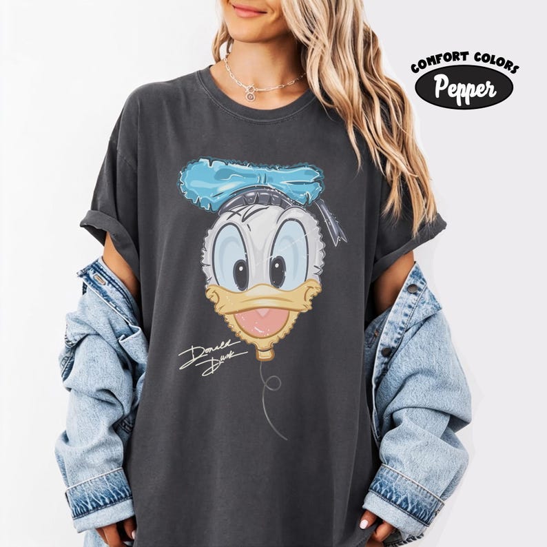 Pode incluir: Uma t-shirt de manga curta cinzento-carv&atilde;o com um gr&aacute;fico do rosto do Pato Donald com um la&ccedil;o azul, um bico amarelo e a assinatura Donald Duck escrita em branco. A t-shirt inclui uma etiqueta Comfort Colors Pepper no canto.