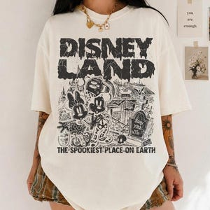 May include: Off-white t-shirt with a black graphic. The design includes the text "DISNEY LAND" and a spooky illustration of Mickey and Minnie Mouse in a graveyard. The text "THE SPOOKIEST PLACE ON EARTH" is also present.
