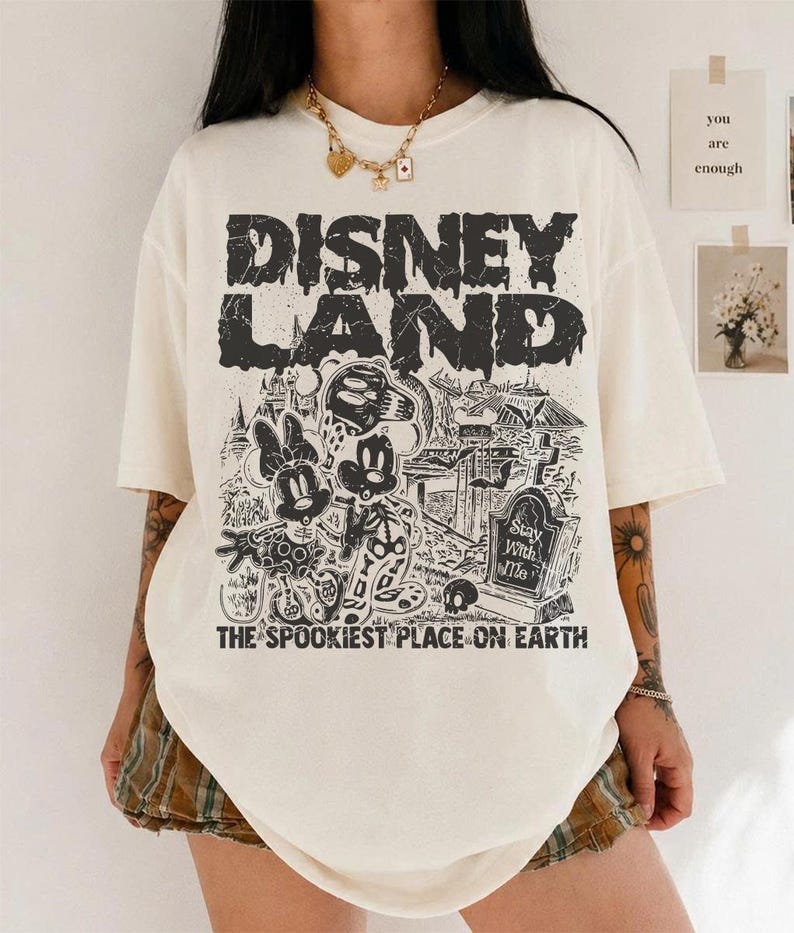 May include: Off-white t-shirt with a black graphic. The design includes the text "DISNEY LAND" and a spooky illustration of Mickey and Minnie Mouse in a graveyard. The text "THE SPOOKIEST PLACE ON EARTH" is also present.