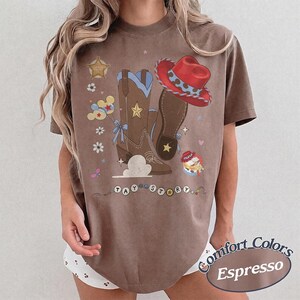 Pu&ograve; includere: Una t-shirt marrone a maniche corte con una stampa grafica di stivali da cowboy marroni, un cappello da cowboy rosso, una stella da sceriffo, palloncini e fiori. Il testo TAY STORY &egrave; stampato in cerchi bianchi in fondo al disegno.