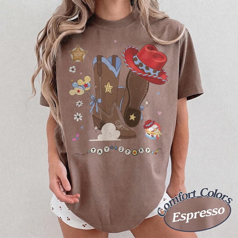 Pu&ograve; includere: Una t-shirt marrone a maniche corte con una stampa grafica di stivali da cowboy marroni, un cappello da cowboy rosso, una stella da sceriffo, palloncini e fiori. Il testo TAY STORY &egrave; stampato in cerchi bianchi in fondo al disegno.