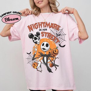 Pu&ograve; includere: Una t-shirt rosa chiaro con una stampa grafica che riporta il testo "NIGHTMARE ON MAIN STREET". Il design include un personaggio scheletro, un cane fantasma, zucche, ragnatele, pipistrelli e un teschio con orecchie da topo su uno sfondo di luna arancione.