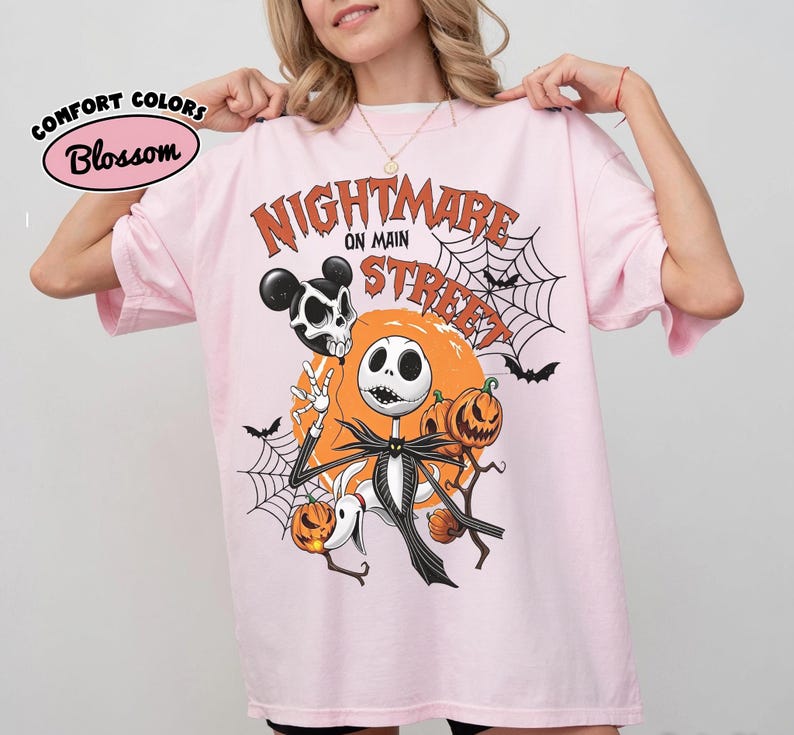 Pu&ograve; includere: Una t-shirt rosa chiaro con una stampa grafica che riporta il testo "NIGHTMARE ON MAIN STREET". Il design include un personaggio scheletro, un cane fantasma, zucche, ragnatele, pipistrelli e un teschio con orecchie da topo su uno sfondo di luna arancione.