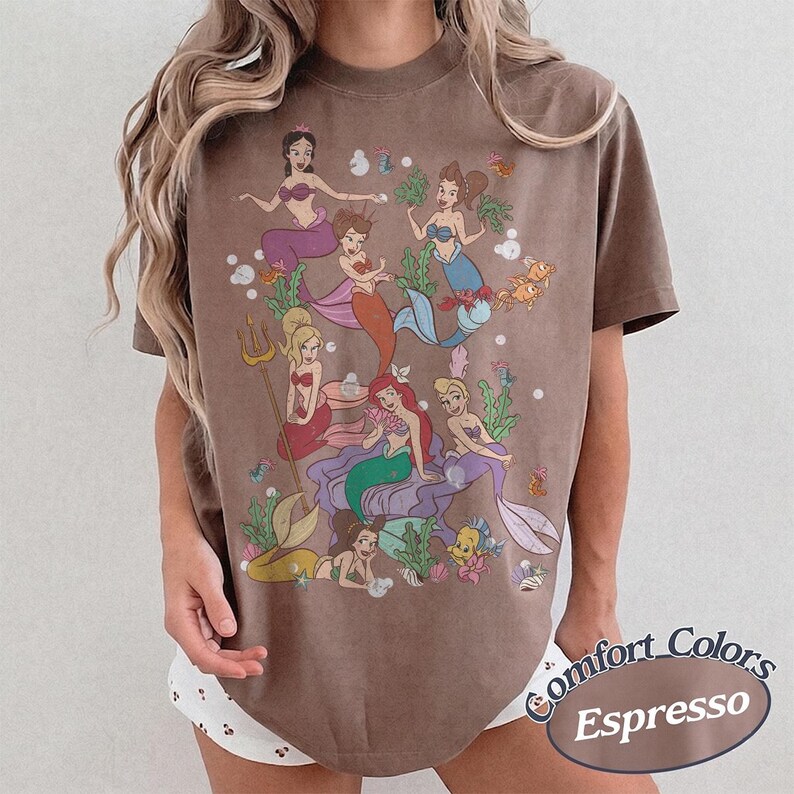 Puede incluir: Una camiseta marr&oacute;n de manga corta con un estampado gr&aacute;fico de varias sirenas con diversos colores de pelo y dise&ntilde;os de cola, rodeadas de elementos submarinos como algas, burbujas y peces peque&ntilde;os. La camiseta tiene cuello redondo.