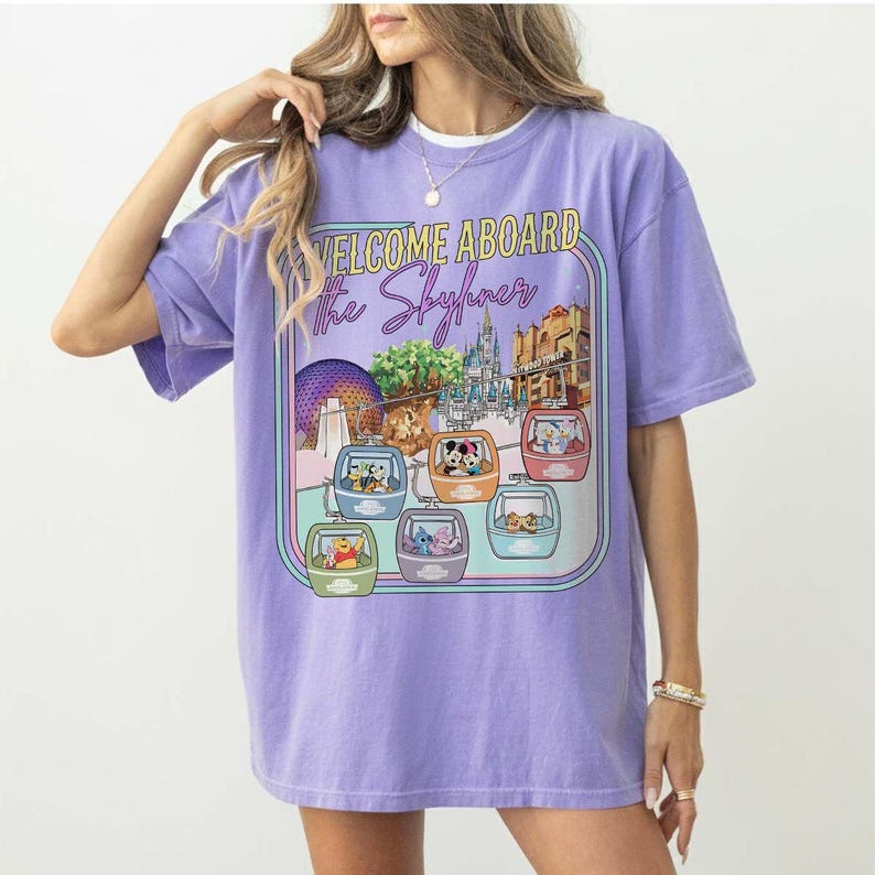 May include: A lilac-colored t-shirt featuring a graphic of a theme park with gondolas and the text "WELCOME ABOARD the Skyliner." The graphic includes cartoon characters and architectural elements.