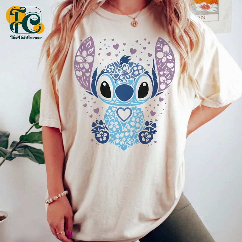May include: A white t-shirt with a blue and purple floral Stitch design. The design features a blue Stitch character with a heart in its chest, surrounded by purple flowers and hearts.