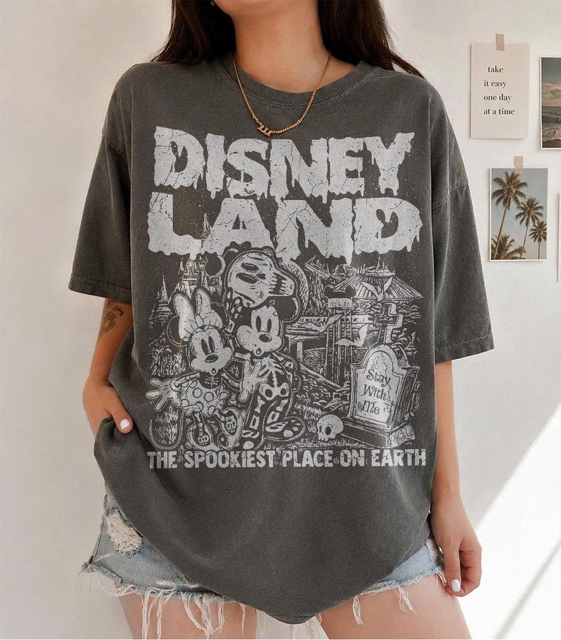 May include: Charcoal grey t-shirt with a distressed "DISNEY LAND" graphic. The graphic features Mickey and Minnie Mouse skeletons in front of a spooky scene, with the text "THE SPOOKIEST PLACE ON EARTH".