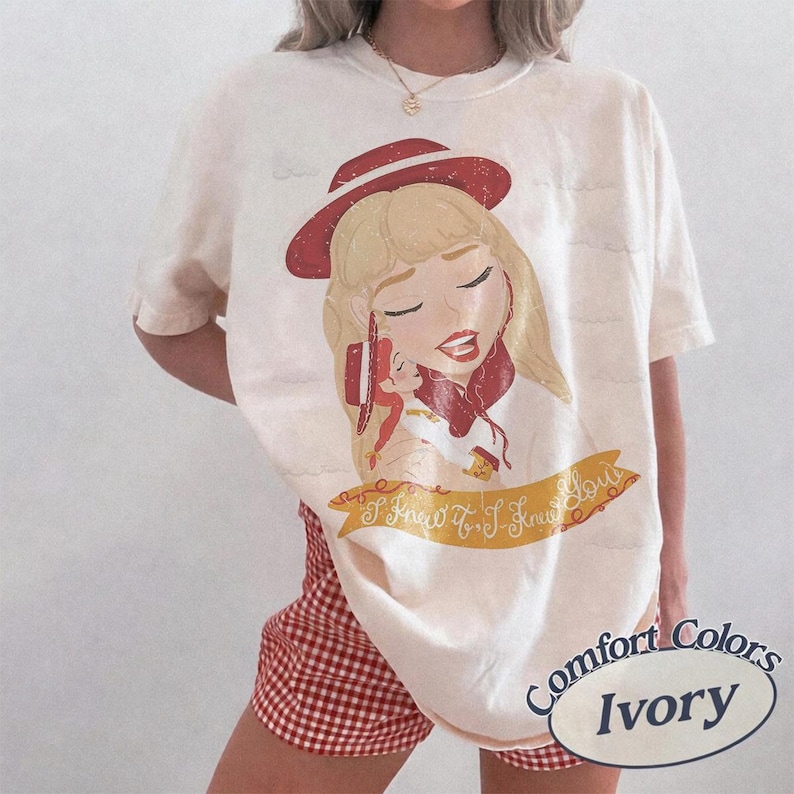 Pode incluir: Uma t-shirt cor de marfim com uma ilustra&ccedil;&atilde;o gr&aacute;fica de uma personagem loira usando um chap&eacute;u e um cachecol vermelhos. Abaixo da personagem, uma faixa amarela exibe o texto em cursivo I Knew It, I Knew You. A t-shirt tem mangas curtas.