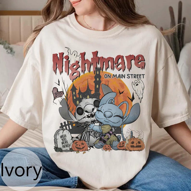 Puede incluir: Camiseta de color marfil con un dise&ntilde;o tem&aacute;tico de Halloween. El dise&ntilde;o incluye la palabra "Nightmare" en rojo, una casa embrujada y personajes de dibujos animados. La camiseta tambi&eacute;n tiene el texto "ON MAIN STREET" y "Wish You Were Here".