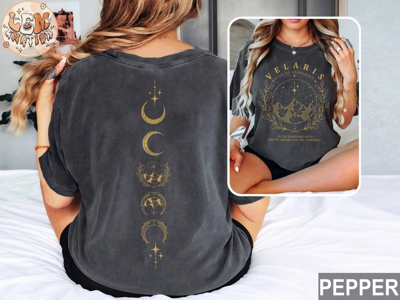 May include: Dark gray t-shirt with a gold design featuring phases of the moon on the back and the text "VELARIS" with a mountain and star design on the front. The shirt is a relaxed fit.