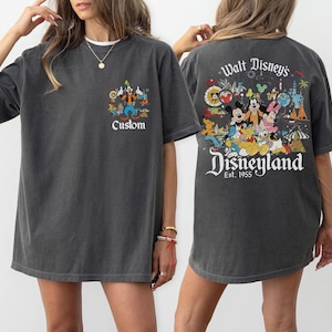 Puede incluir: Camiseta gris oscuro con un gr&aacute;fico de Disneyland en la parte delantera y trasera. El gr&aacute;fico frontal incluye la palabra "Custom". El gr&aacute;fico trasero dice "Walt Disney's Disneyland Est. 1955" con personajes de dibujos animados.