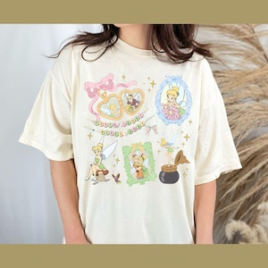 May include: A cream colored t-shirt with a graphic print of Tinkerbell and other fairies. The print features multiple frames with Tinkerbell in different poses, including flying, sitting on a mushroom, and holding a jar of pixie dust. The text "Faith, Trust, Pixie Dust" is printed on the shirt.