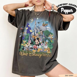 May include: A charcoal gray short-sleeve t-shirt featuring a graphic print of various Disney characters in front of a castle, with the text Walt Disney World. The shirt has a crew neckline and a loose fit, with Comfort Colors Pepper branding.