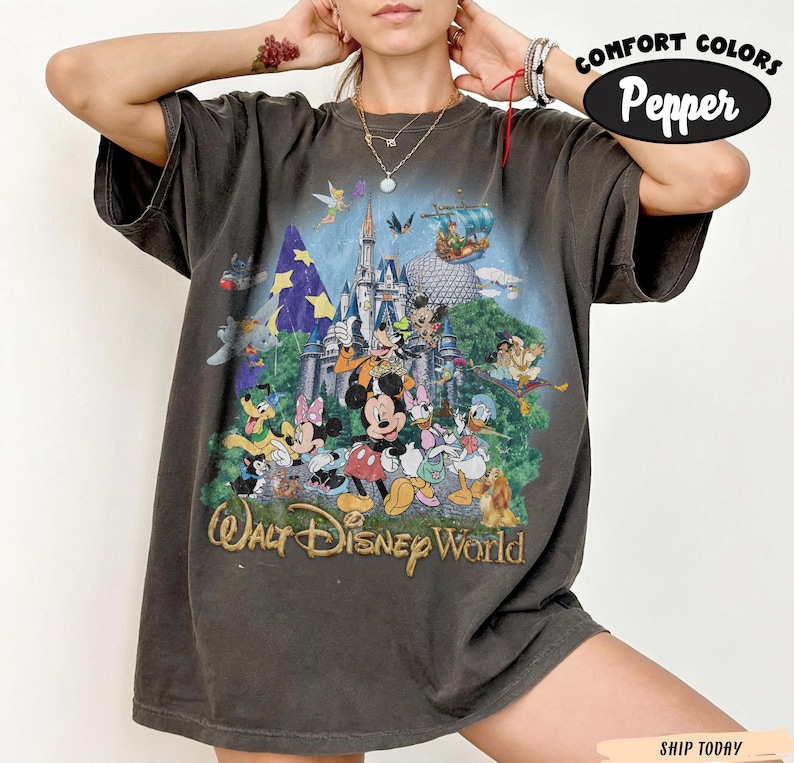 May include: A charcoal gray short-sleeve t-shirt featuring a graphic print of various Disney characters in front of a castle, with the text Walt Disney World. The shirt has a crew neckline and a loose fit, with Comfort Colors Pepper branding.