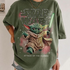 K&ouml;nnte beinhalten: Olivgr&uuml;nes T-Shirt mit einer Grafik von Yoda aus Star Wars, der ein gr&uuml;nes Lichtschwert h&auml;lt. Der Text "STAR WARS" steht &uuml;ber der Figur und "ATTACK OF THE CLONES" darunter. Das T-Shirt hat kurze &Auml;rmel.