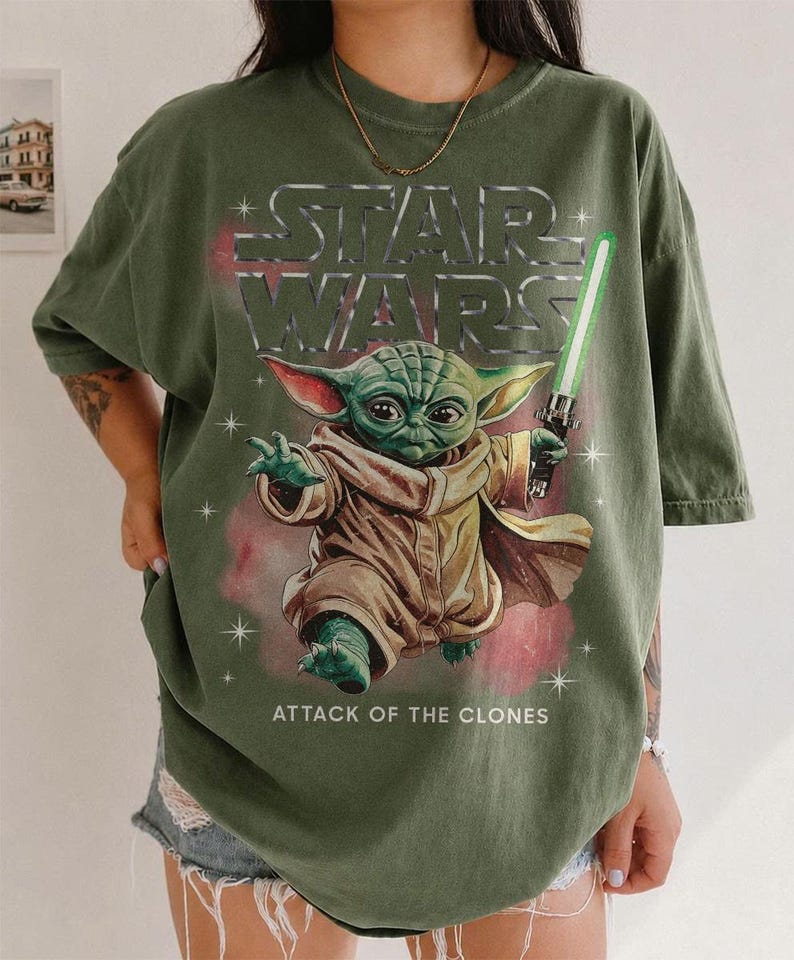 K&ouml;nnte beinhalten: Olivgr&uuml;nes T-Shirt mit einer Grafik von Yoda aus Star Wars, der ein gr&uuml;nes Lichtschwert h&auml;lt. Der Text "STAR WARS" steht &uuml;ber der Figur und "ATTACK OF THE CLONES" darunter. Das T-Shirt hat kurze &Auml;rmel.