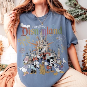May include: Blue t-shirt featuring a colorful graphic of Disneyland's castle and various Disney characters, including Mickey Mouse, Minnie Mouse, and Donald Duck. The shirt has the text "Walt Disney Disneyland" in a rainbow font.