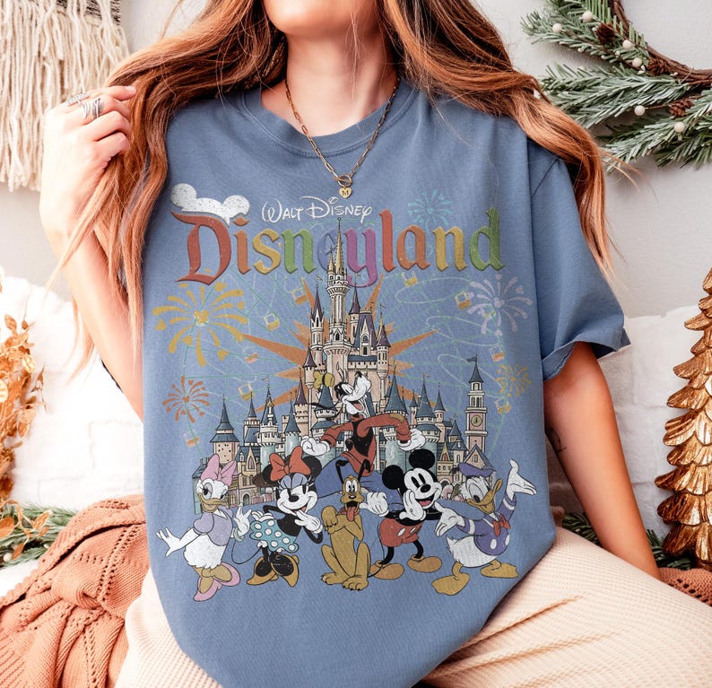 May include: Blue t-shirt featuring a colorful graphic of Disneyland's castle and various Disney characters, including Mickey Mouse, Minnie Mouse, and Donald Duck. The shirt has the text "Walt Disney Disneyland" in a rainbow font.