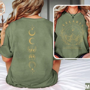 May include: Moss green t-shirt with a gold moon phase design on the back and a framed graphic on the front. The front graphic reads "VELARIS CITY OF STARLIGHTS" with a mountain and star design. The text "THE STARS WHO LISTEN AND THE DREAMS THAT ARE ANCHOR" is also included.