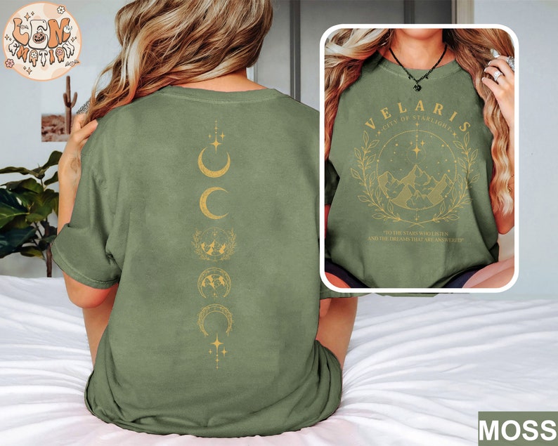 May include: Moss green t-shirt with a gold moon phase design on the back and a framed graphic on the front. The front graphic reads "VELARIS CITY OF STARLIGHTS" with a mountain and star design. The text "THE STARS WHO LISTEN AND THE DREAMS THAT ARE ANCHOR" is also included.