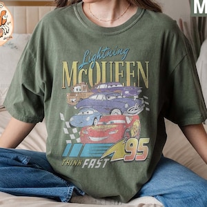 May include: A green t-shirt with a vintage-style graphic of Lightning McQueen from the Cars movie. The graphic features McQueen and other cars from the movie, with the text "Lightning McQueen" and "Think Fast" printed on the shirt.